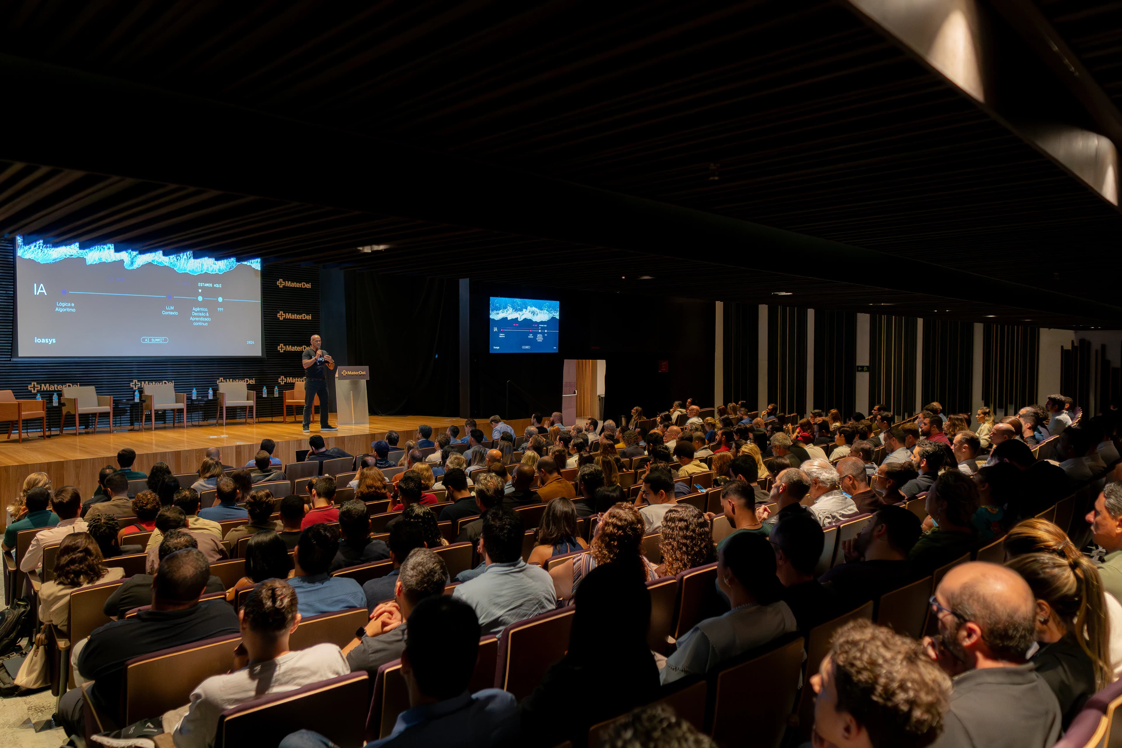 Auditório lotado acompanhando uma palestra do AI Summit, com o palestrante ao púlpito e o slide projetado ao fundo